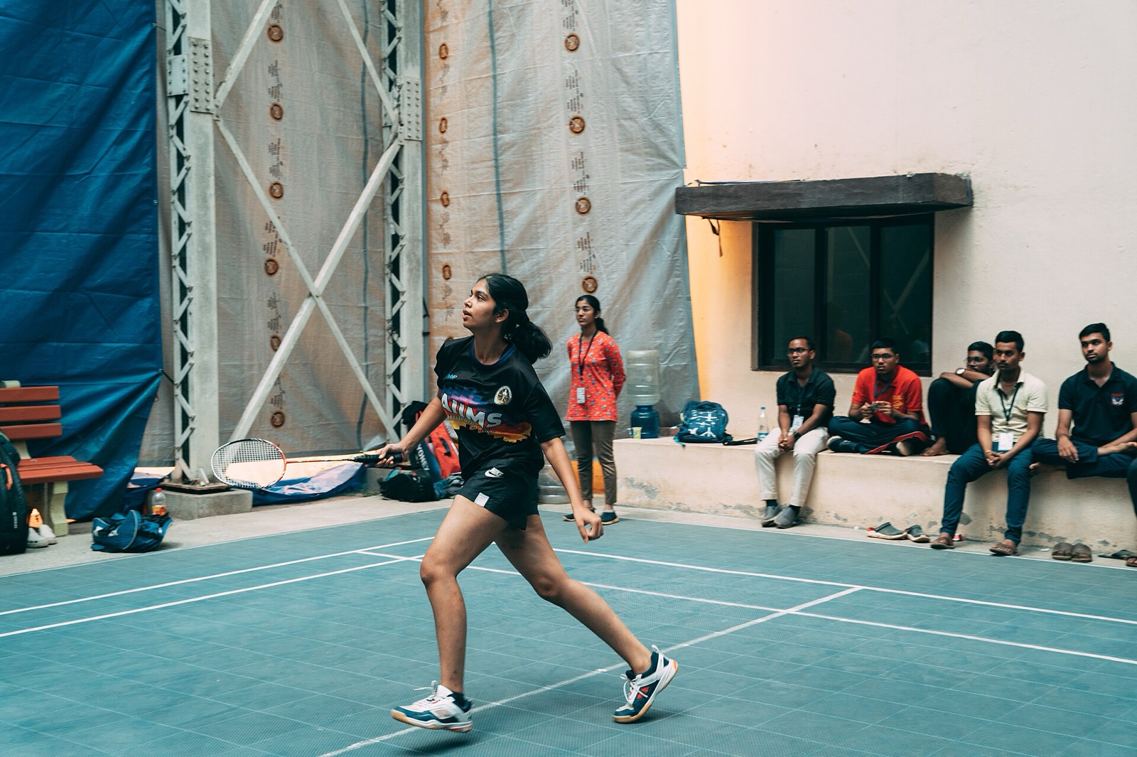A badminton player stretching to hit a shuttle on an indoor court in India while spectators sit along the wall