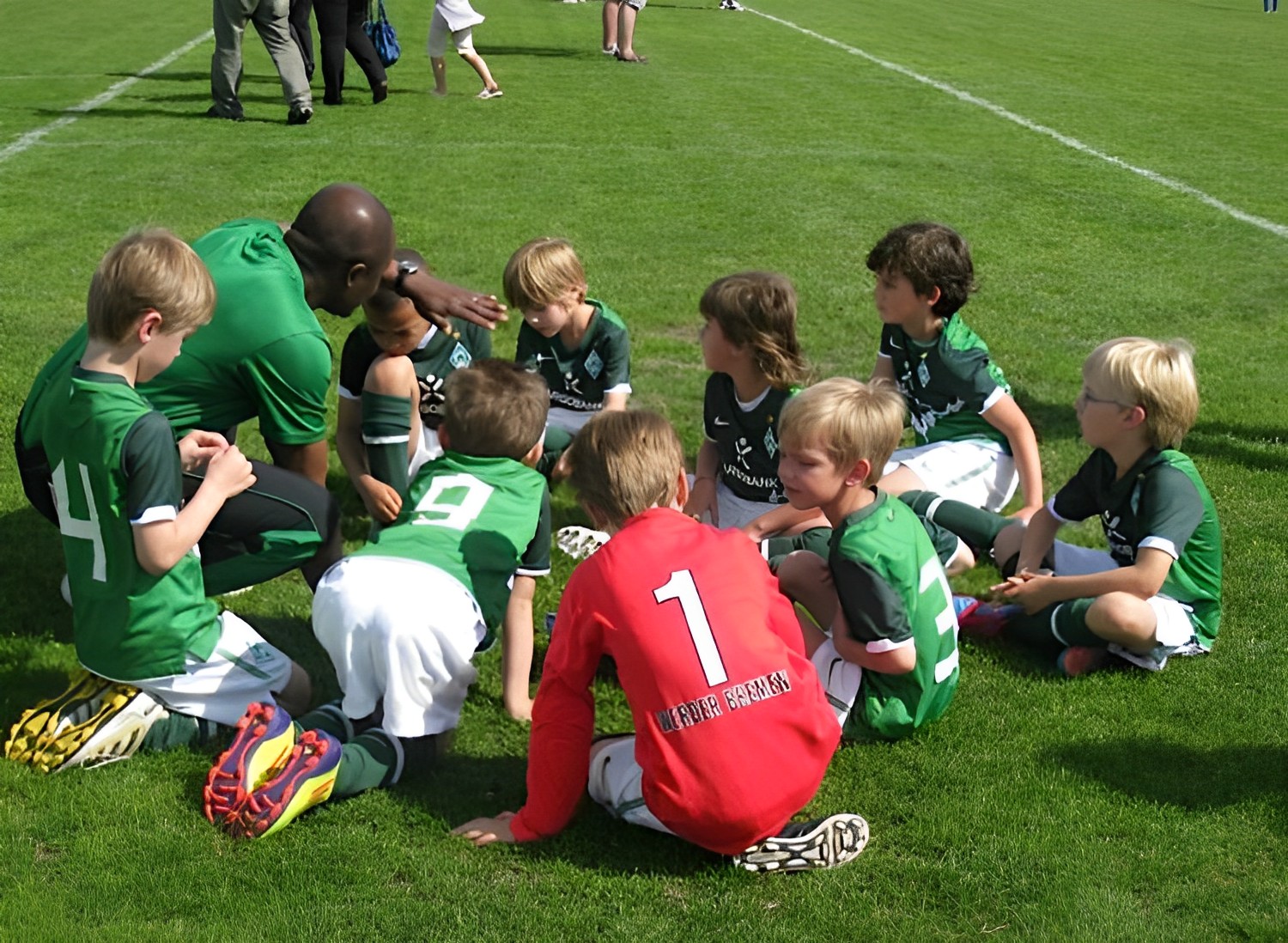 A youth football coach talking with a group of young players sitting on the pitch